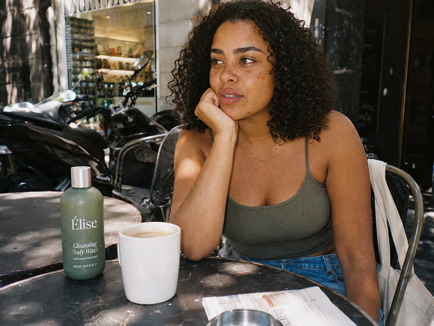 A woman at a street café beside one Elise body-wash bottle and a white coffee cup