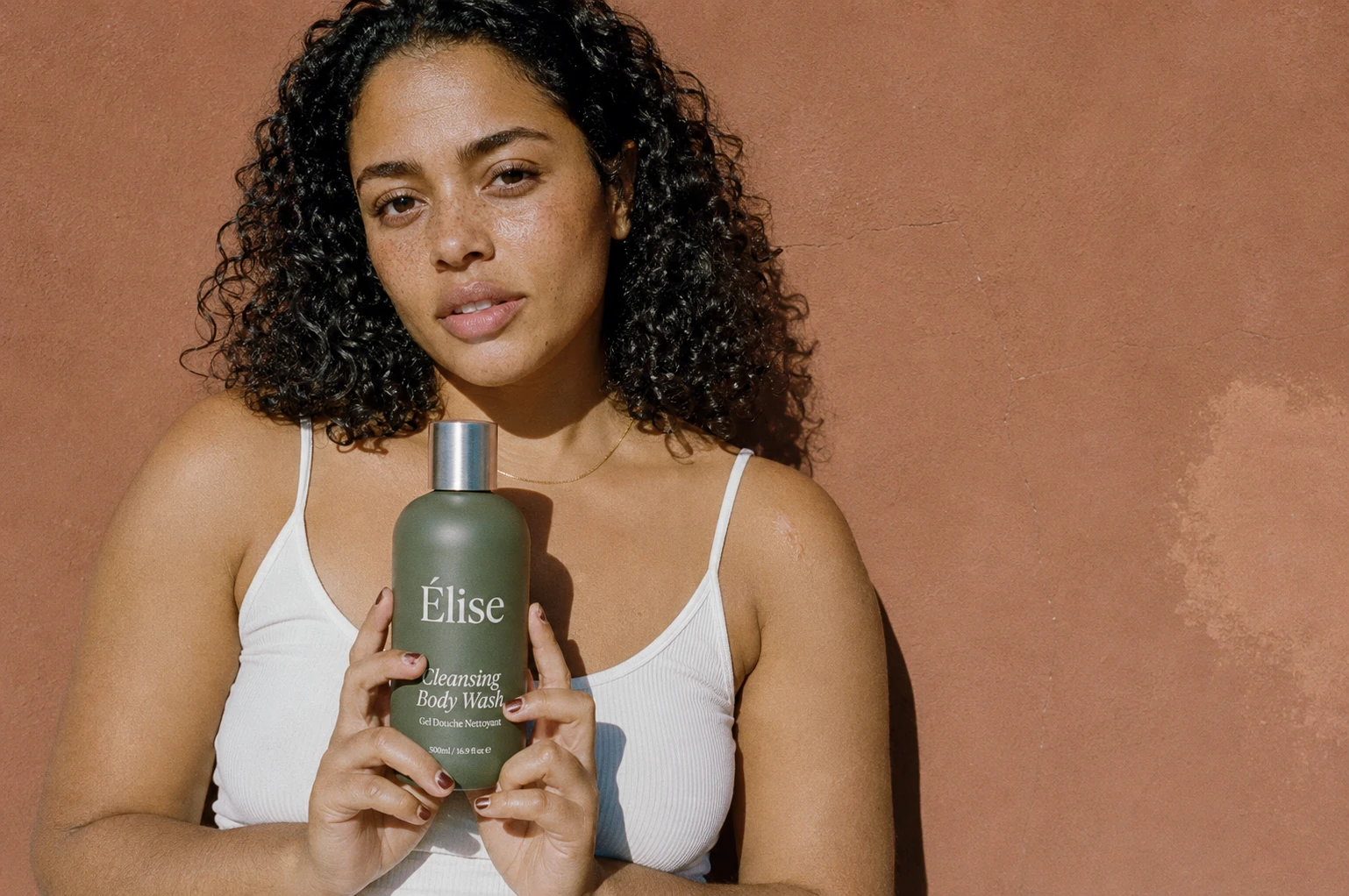 A woman holding one Elise body-wash bottle against a warm terracotta wall