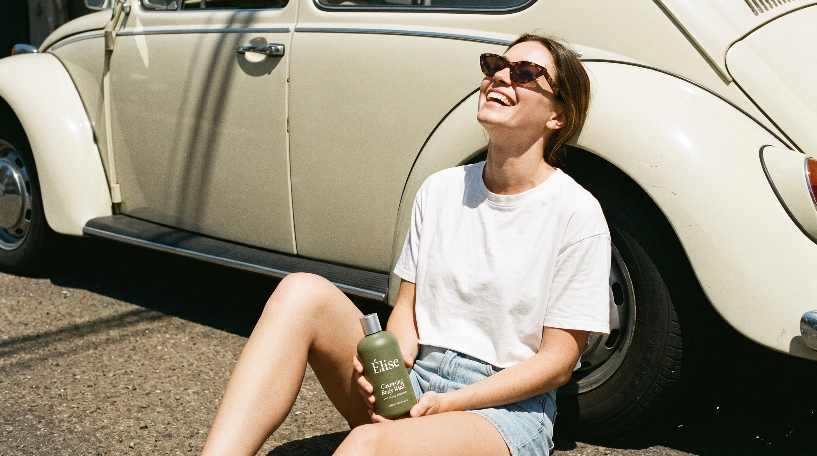 A laughing woman seated beside a vintage cream Volkswagen while holding one Elise body-wash bottle
