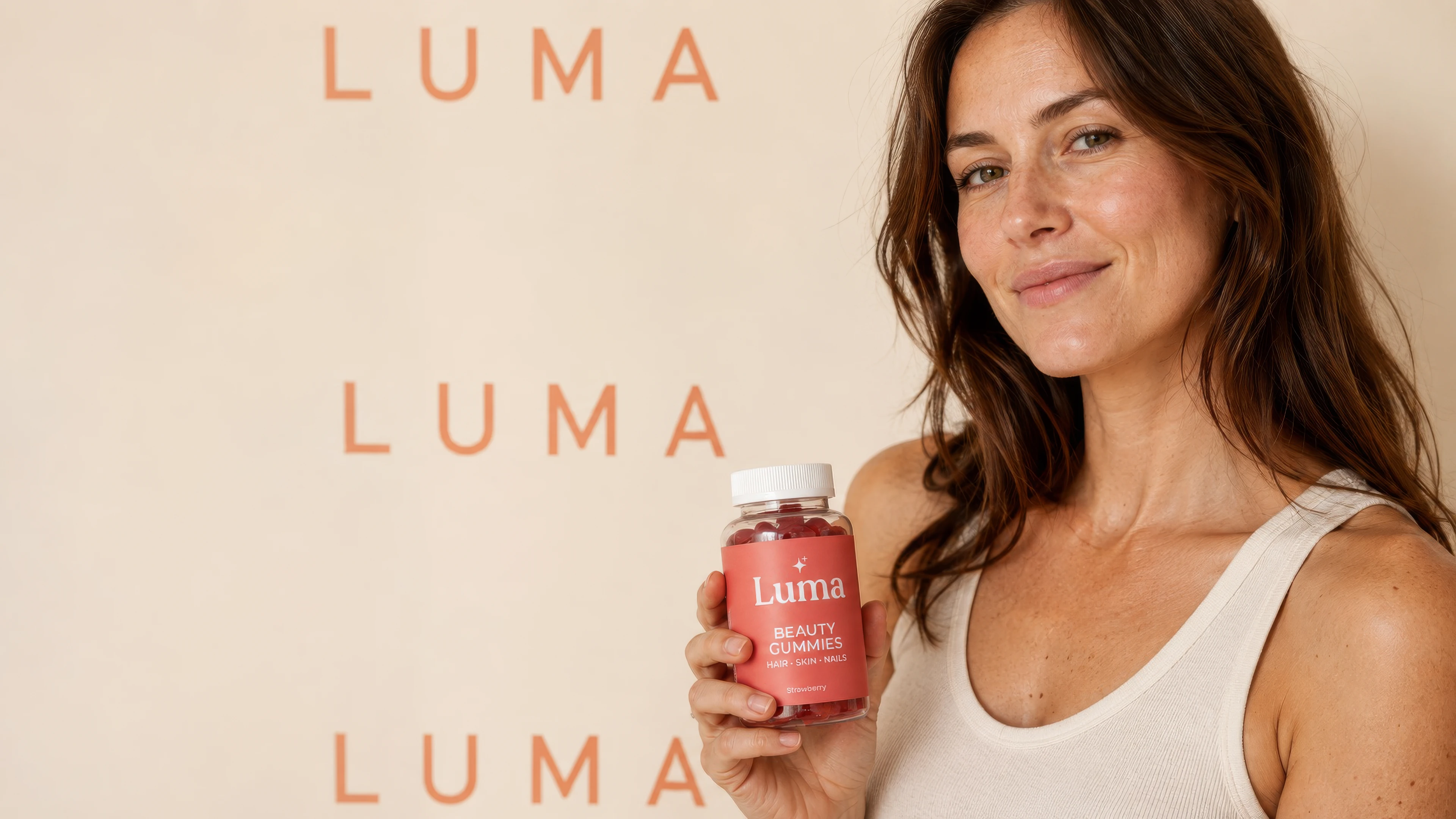A professional white female model naturally holding a realistic strawberry Luma beauty-gummies jar against a repeated LUMA wordmark backdrop