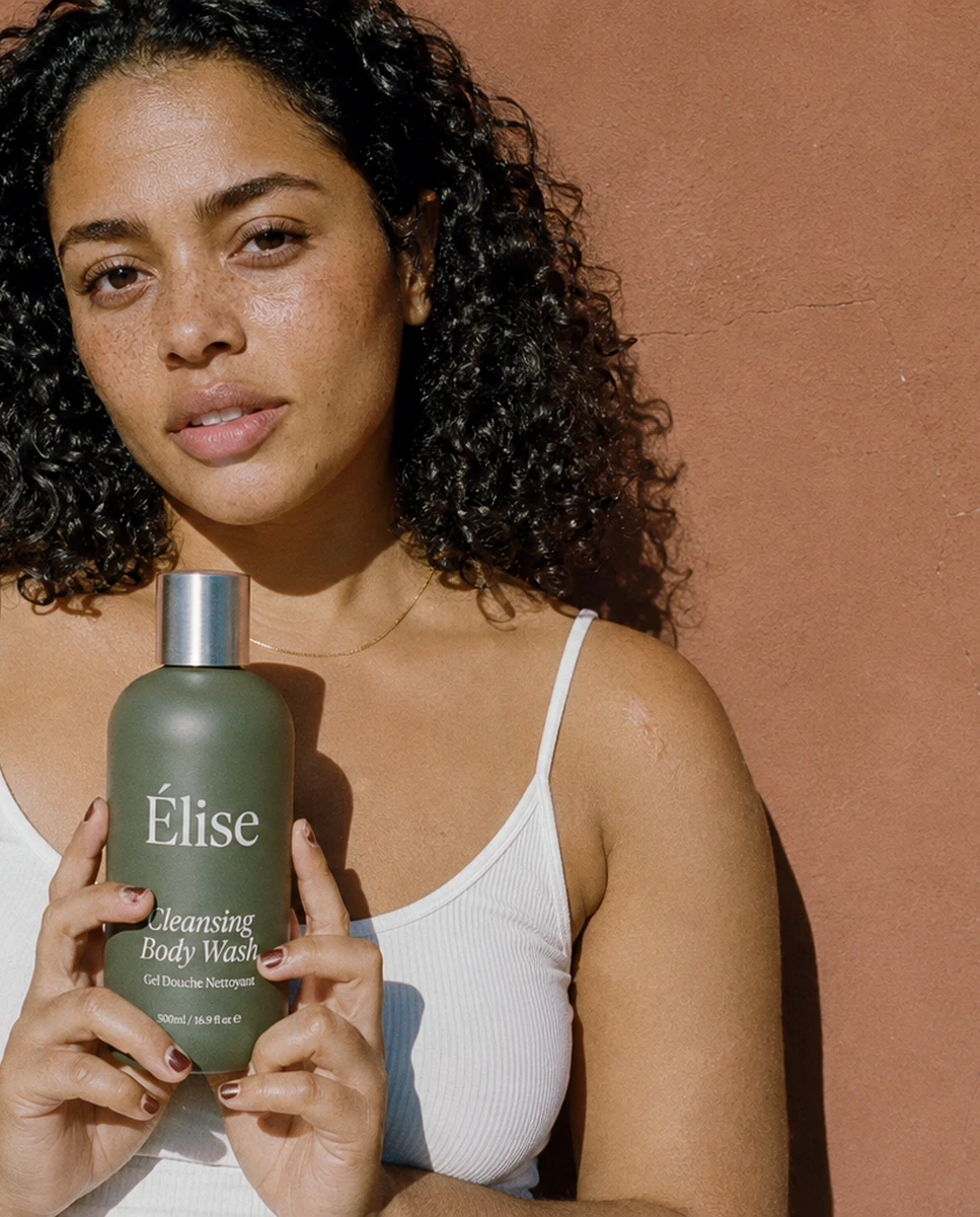 A woman holding one Elise body-wash bottle against a warm terracotta wall
